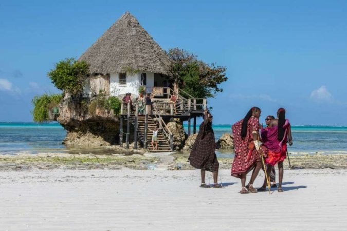 Des locaux en tenue traditionnelle discutent sur une plage près d'un restaurant sur un rocher.